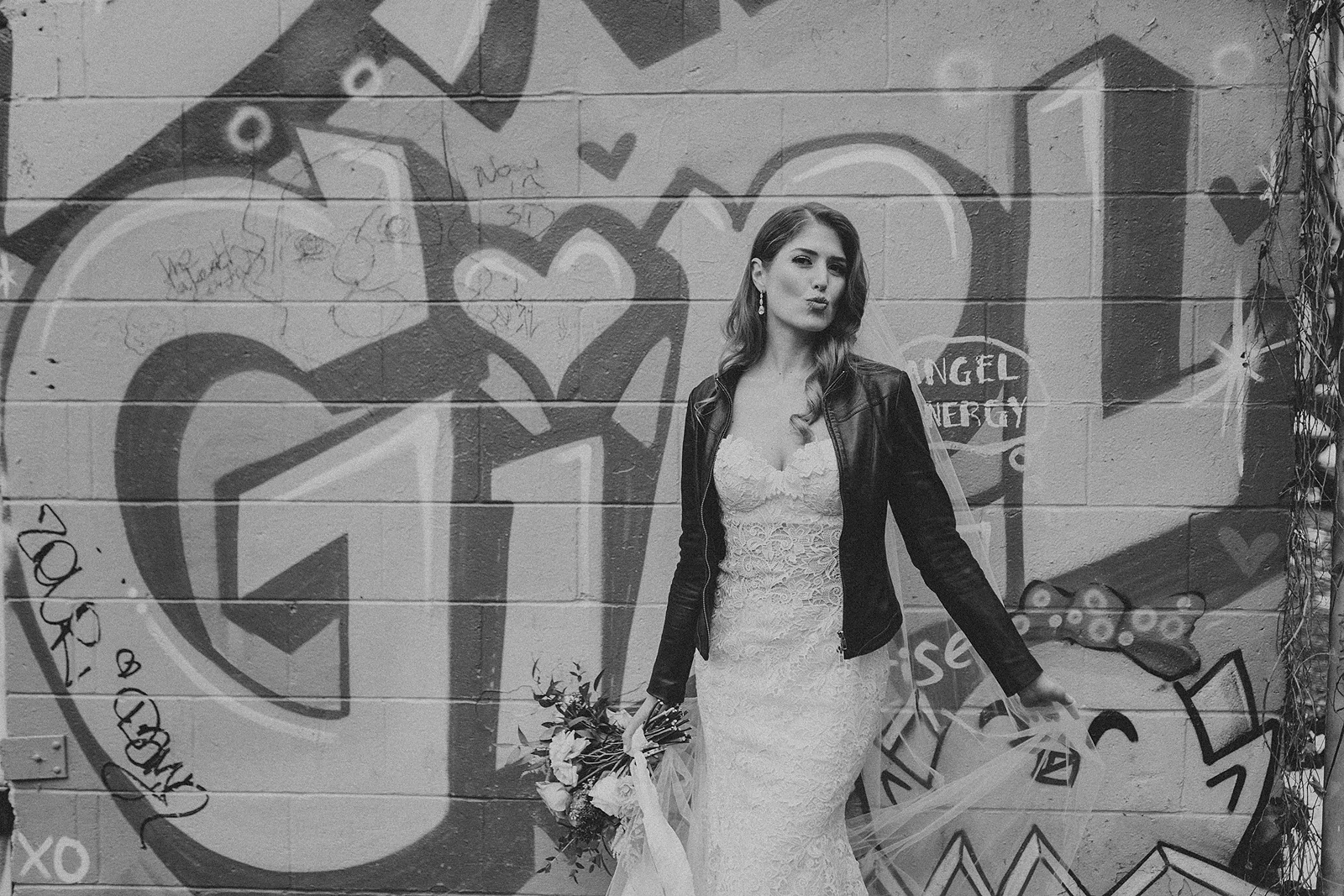 a bride wearing a leather jacket over her wedding dress standing infront of a graffiti covered wall that reads "Girl"