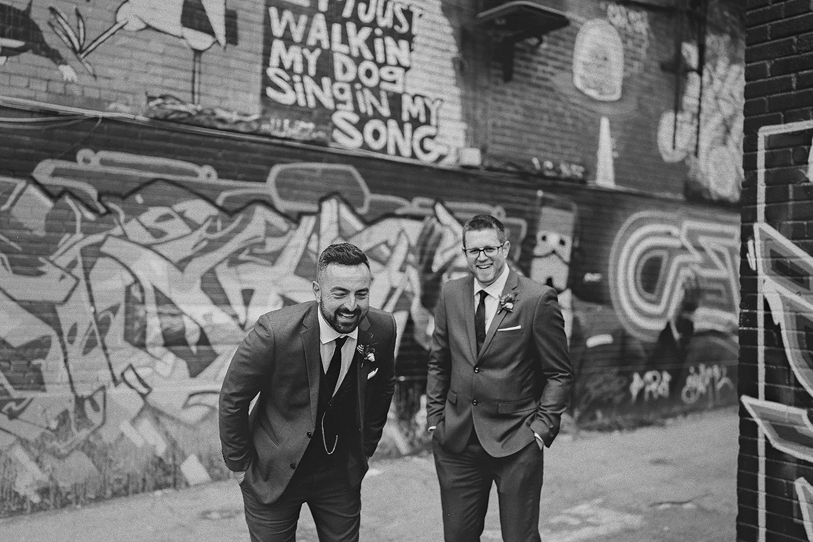 Two groomsmen laugh in front of a graffiti wall