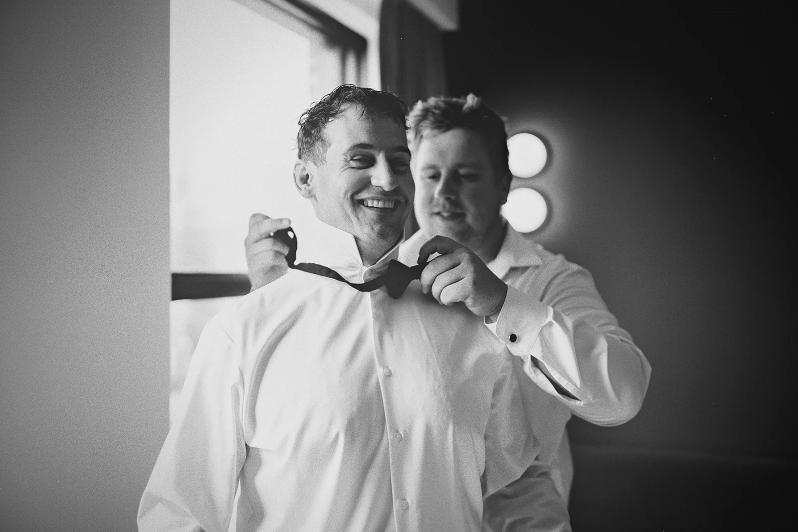 A groomsman helps a smiling Kieran with his bow tie