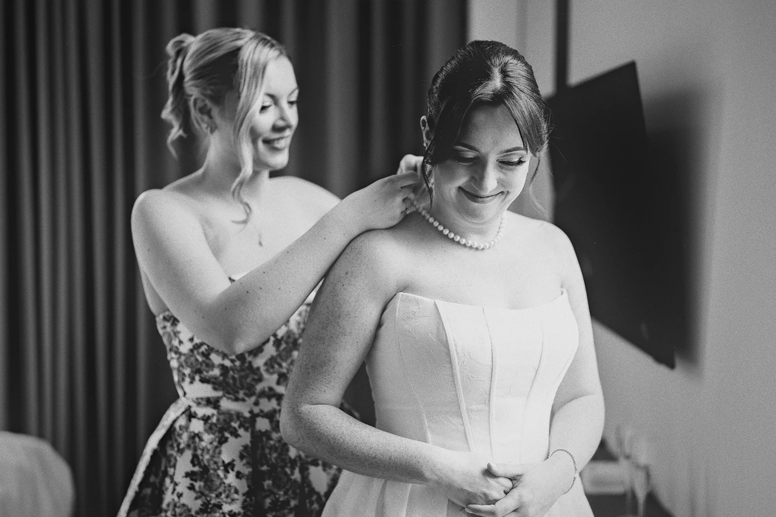 Hillary smiles as her bridesmaid puts on her necklace.