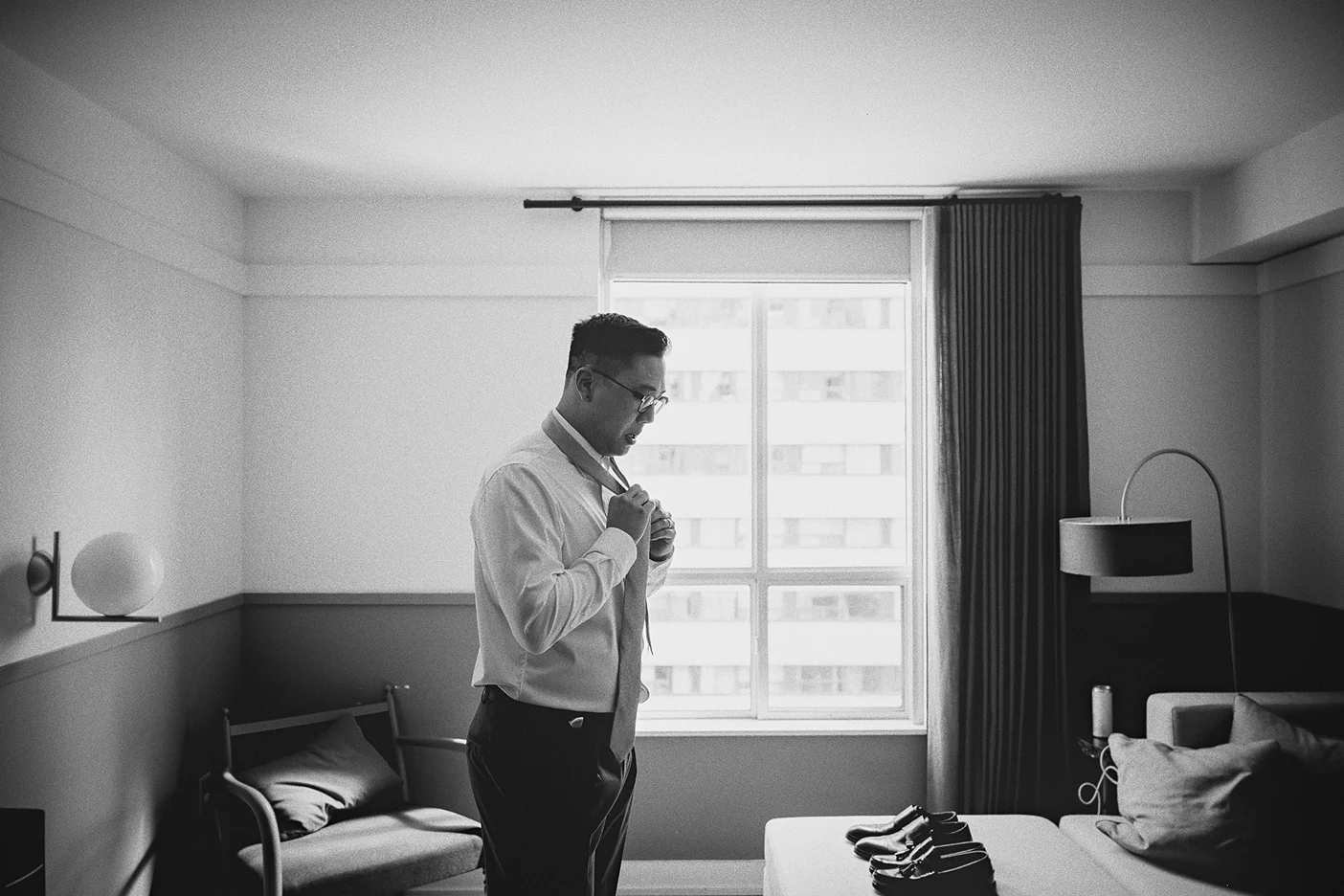 Ky is silhouetted against a bright window as he adjusts his light grey tie.