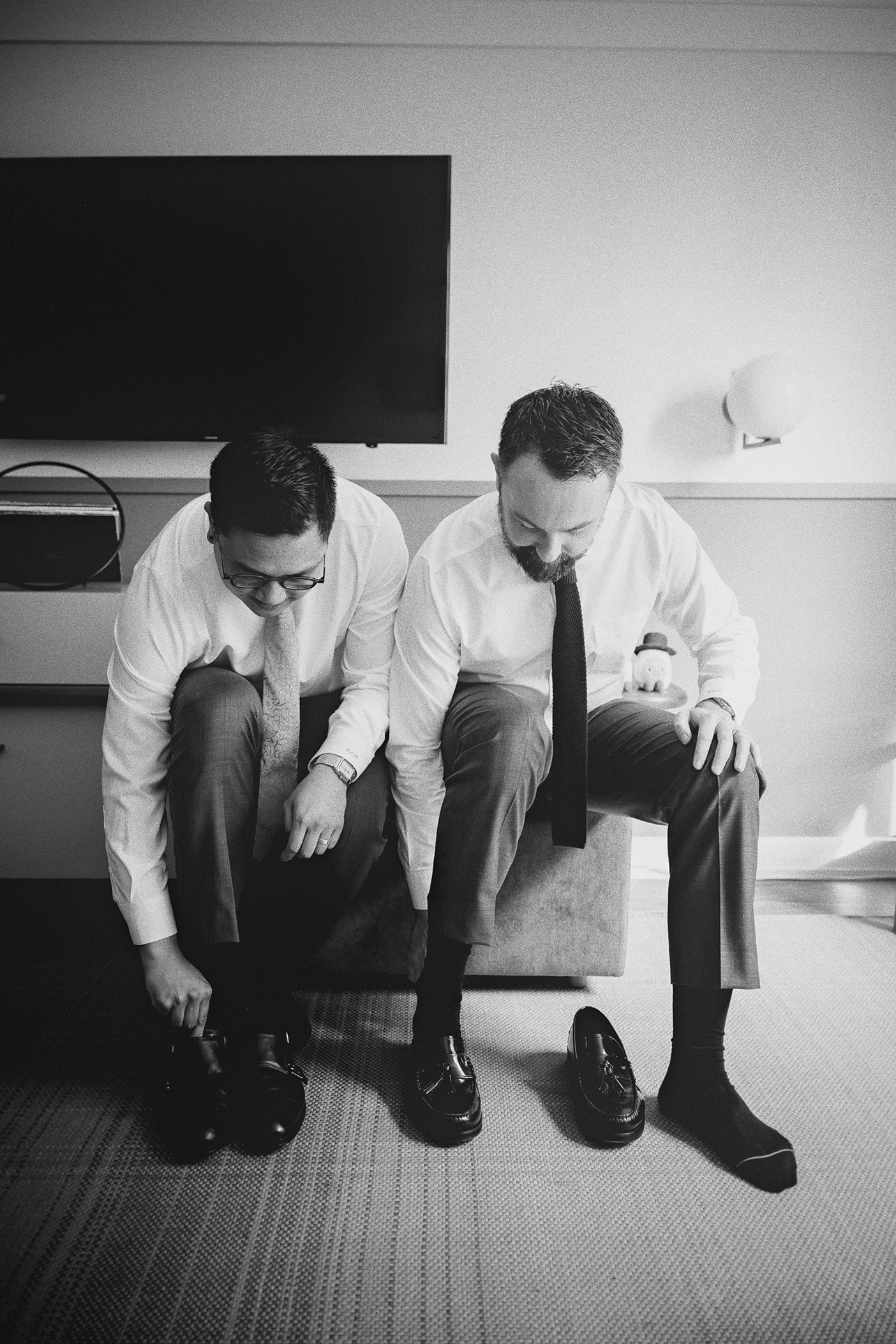 The grooms sit side-by-side, each focused on putting on his wedding shoes.