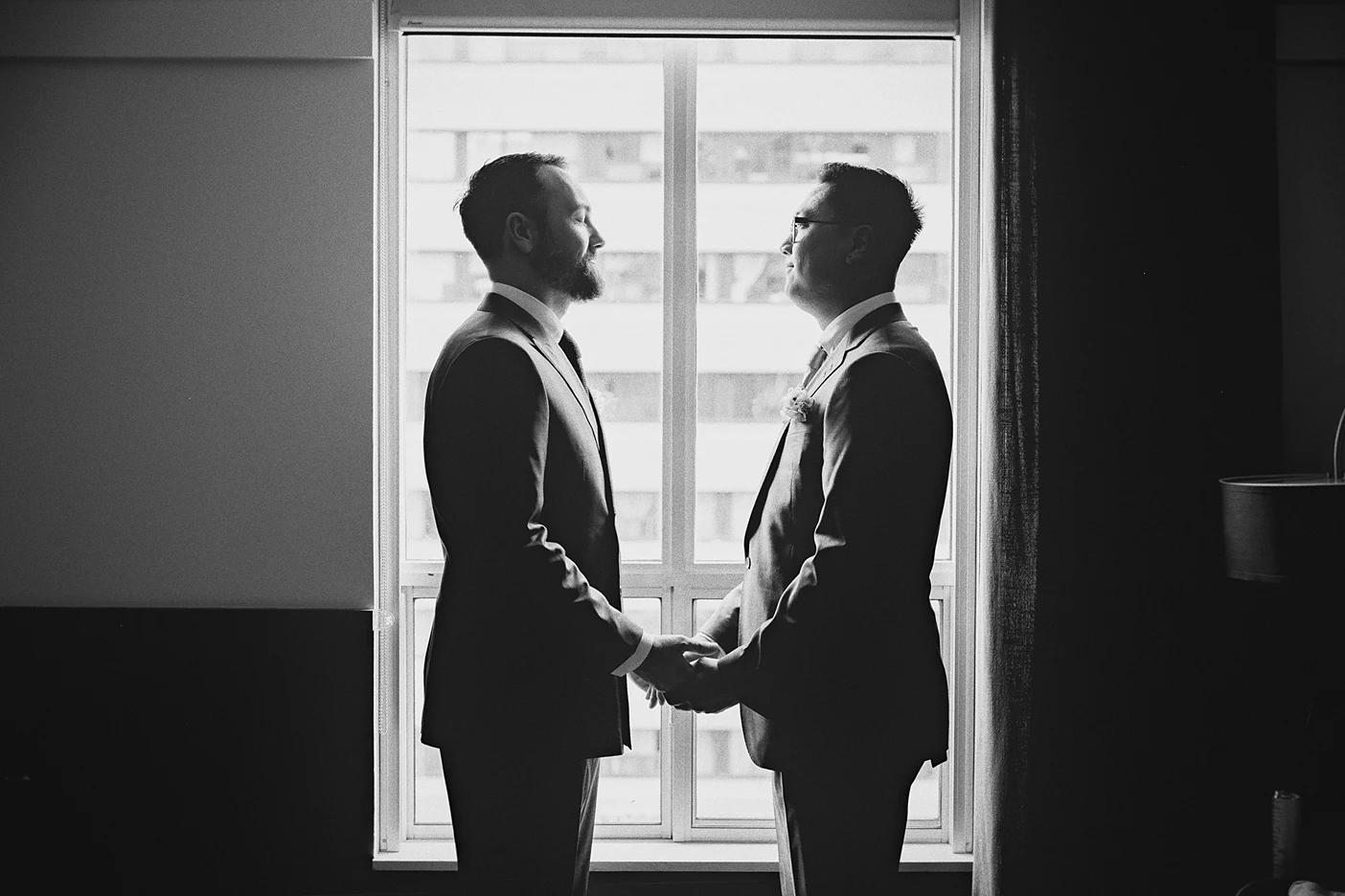 Silhouetted against the window, the grooms hold hands in a quiet, intimate moment.