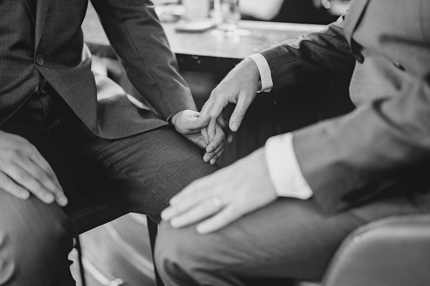 A close-up shows the grooms' hands clasped together as they sit at the bar.