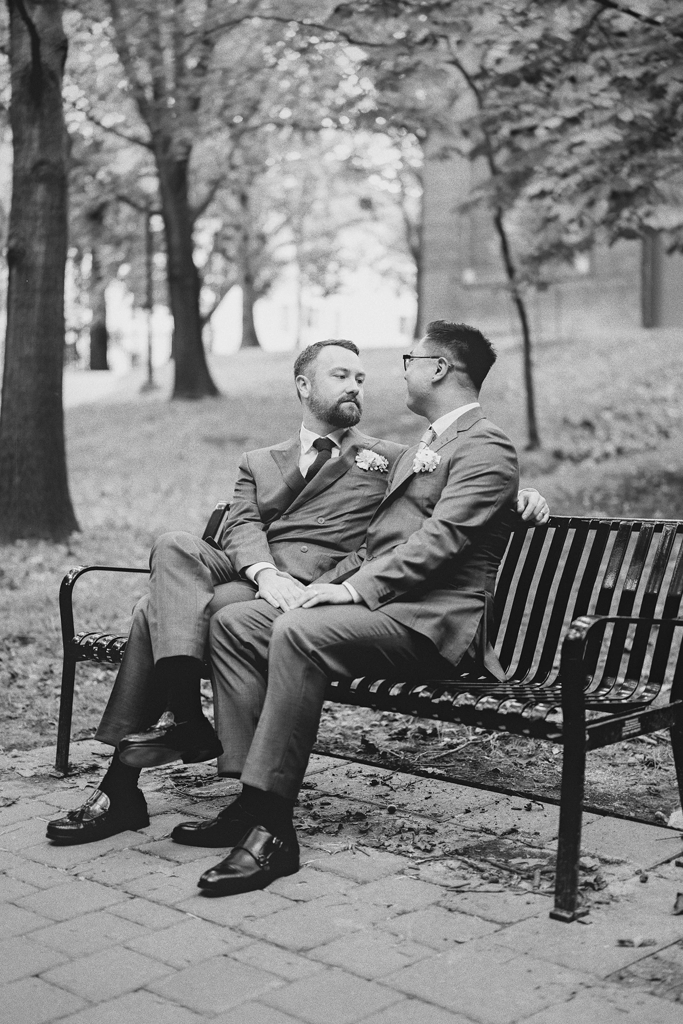 The grooms sit closely together on a park bench, looking at each other.