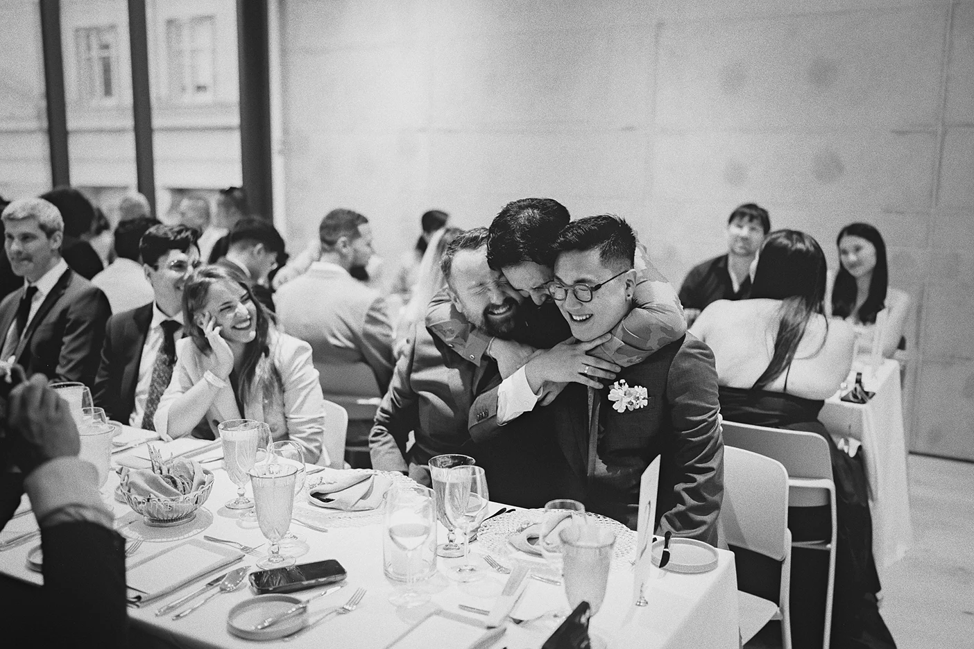 A guest embraces both grooms at their dinner table.