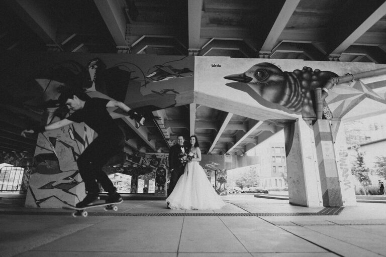 A bride and groom stand together under an overpass in Toronto and watch a skateboarder skating past